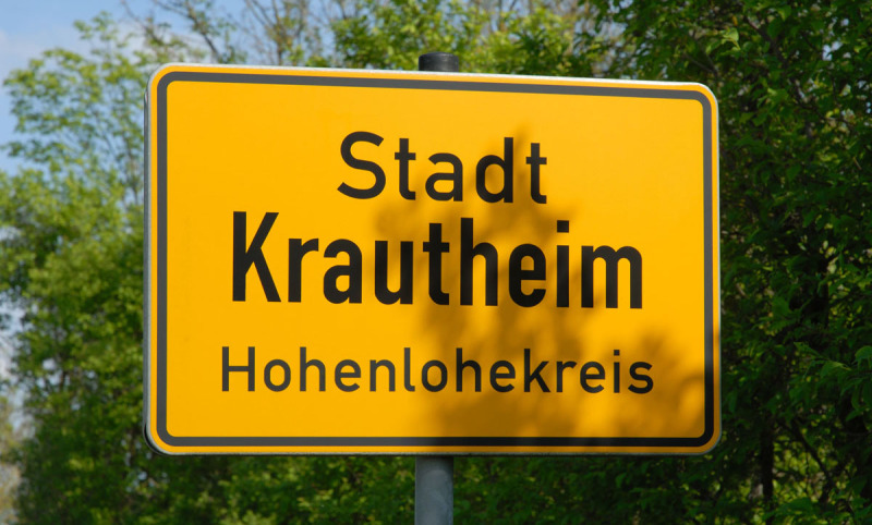 Das Orsteingangsschild von Krautheim, Hohenlohe ist gelb mit schwarzer Schrift. Rund herum ist grünes Laub von Bäumen. Hier sucht die Spedition Lkw-Fahrer.