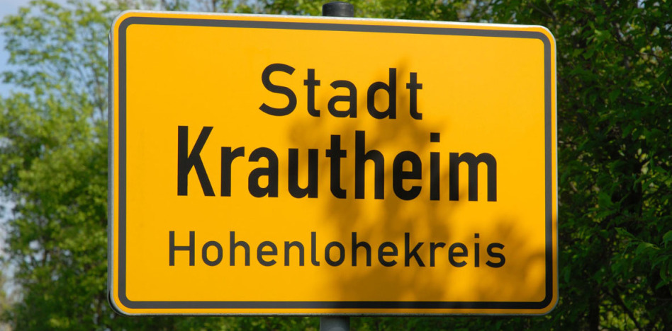Bildfüllend ist das Ortsschild der Stadt in Baden-Württemberg zu sehen. Im Hintergrund ist hellgrünes Frühlingslaub. Auf dem gelben Schild steht Stadt Krautheim, Hohenlohekreis.