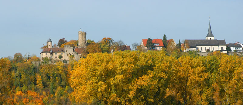 Man sieht Krautheim in Baden-Württemberg von unten, im unteren Drittel sind herbstliche Bäume, im oberen Drittel Himmel, dazwischen sind die Burg und die Kirche.