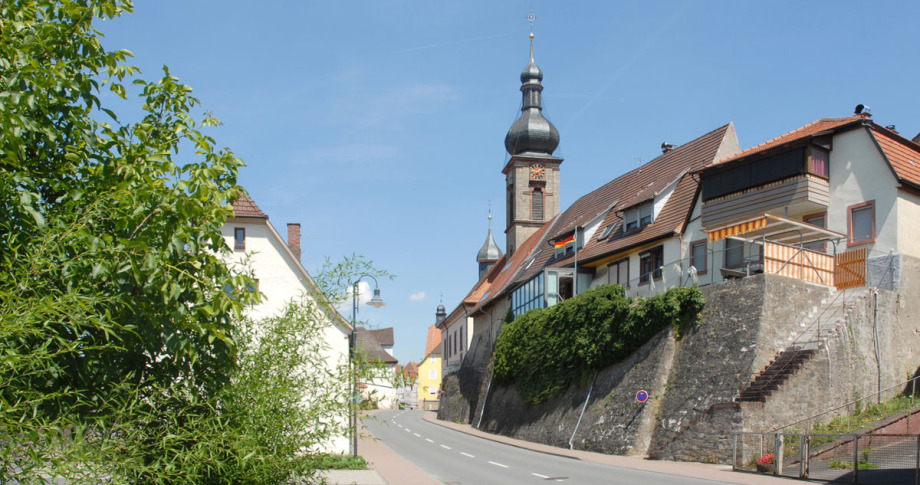 Eine Straße in Boxberg führt wie in einer kleinen Schlucht zur Bildmitte. Auf der rechten Anhöhe stehen vorne Wohnhäuser, hinten sieht man den Turm einer Kirche.