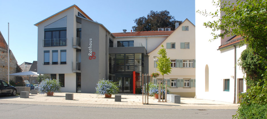 Das Rathaus in Dörzbach mit Vorplatz. Es ist ein moderner Neubau. Rechts davon ist ein älteres Wohnhaus. Davon rechts erahnt man eine Kirche. Der Vorplatz ist mit Blumentrögen geschmückt, darunter verläuft eine Straße.