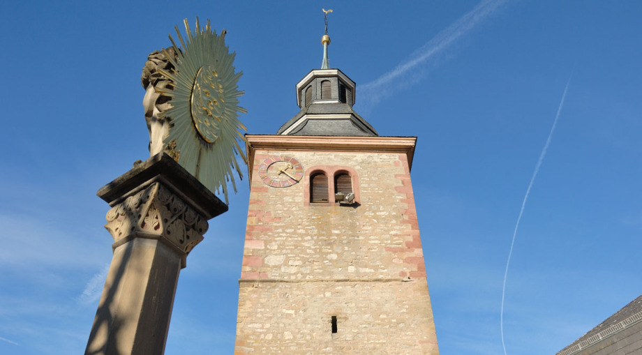 Man sieht den Kirchturm in Osterburken hinauf zur Uhr und zur Spitze. Davor, links im Bild, ist eine Pestsäule, deren oberer Teil ein goldenes Wappen mit Strahlenkranz bildet. Der Himmel ist blau, die Szenerie ist sonnenbeschienen.