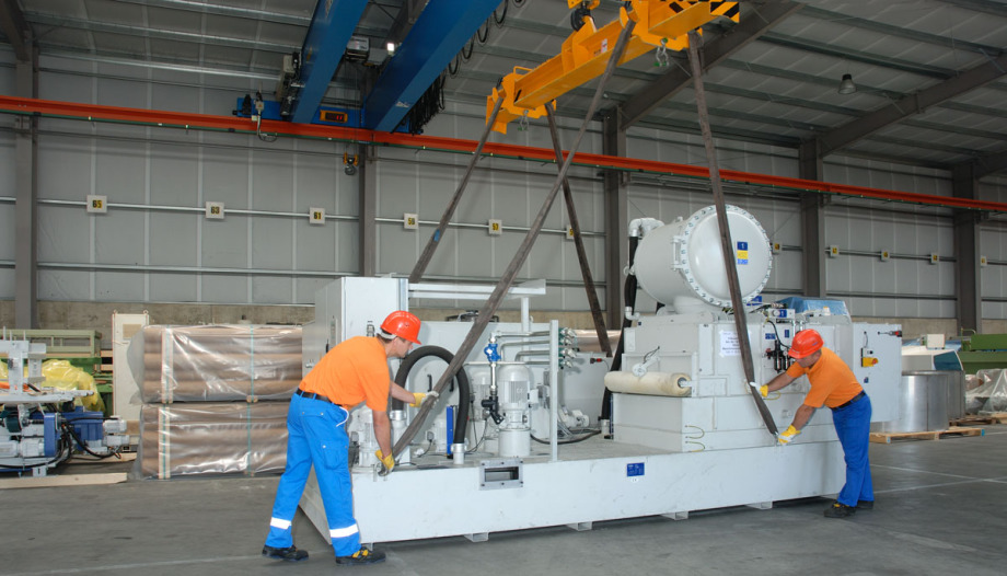 Zwei Mitarbeiter in blau/orangener Montur und Helm arbeiten an einer hellgrauen, großen Maschine, die an Seilen von einem Kran darüber angehoben werden soll. Im Hintergrund sind verpackte Güter.