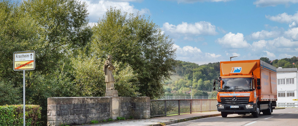 Ein 7,5-Tonnen-Lkw fährt über eine Brücke auf den Betrachter zu. Links ist eine historische Heiligenfigur, davor ein Ortsschild. Hinter der Brücke sieht man grüne Bäume, im Hintergrund des Bildes ist Wald. Rechts sieht man ein weißes Gebäude im Hintergrun