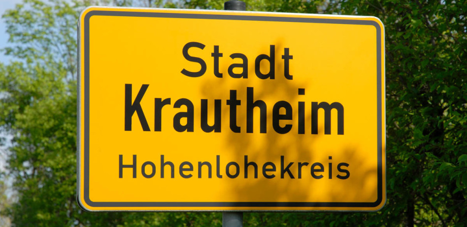 Riesig ist das Ortseingangsschild von Krautheim fotografiert. Dahinter sind Bäume mit hellgrünem Laub. Auf dem Schild steht: Stadt Krautheim ... Hohenlohe.