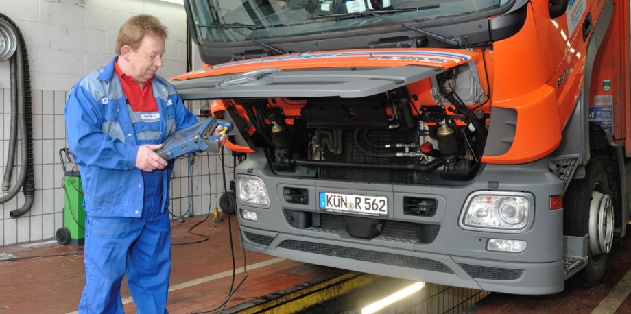 Ein Mitarbeiter mit mobilem elektronischen Gerät steht vor der aufgeklappten Motorhaube eines Lkw. Er hat einen blauen Overall an.