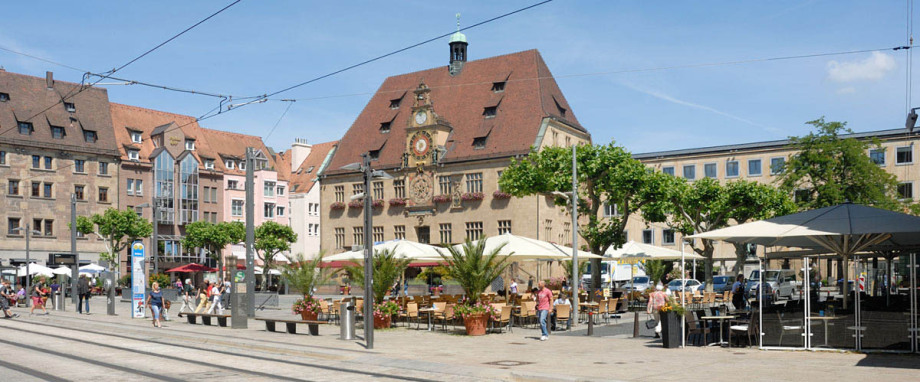 Der Marktplatz von Heilbronn mit dem historischen Rathaus. Vorne rechts stehen aufgeklappte Schirme eines Restaurants. Links vorne sind Straßenbahnschienen sichtbar. Am Rand des Marktplatzes ist eine Häuserzeile.