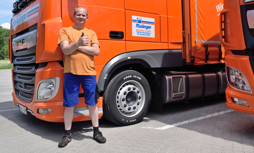 Ein LKW-Fahrer der Spedition in Hohenlohe steht vor seinem orangenen LKW. Es ist ein kurzer LKW. Er hat eine Hand an der Türklinke, mit der anderen schaut er auf ein Handy.