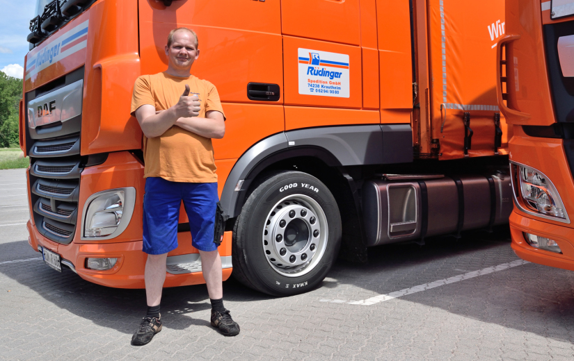 Ein Lkw-Fahrer steht vor dem Fahrerhaus seines Lkws. Er hat ein orangenes T-Shirt an und kurze, blaue Hosen bis zu den Knieen. Der Fahrer zeigt dem Betrachter „den Daumen hoch“. Rechts im Bild sieht man einen winzigen Teil eines weiteren Lkw.