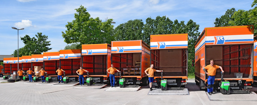 Mehrere 7,5-Tonnen-Lkw sind sorgfältig und sauber, etwas versetzt, aufgereiht und man sieht von der Rückseite in die offenen Fahrzeuge. Die Ladeklappen sind alle auf den Hof abgesenkt und acht Mitarbeiter in orangenem T-Shirt und blauer Hose stehen neben 
