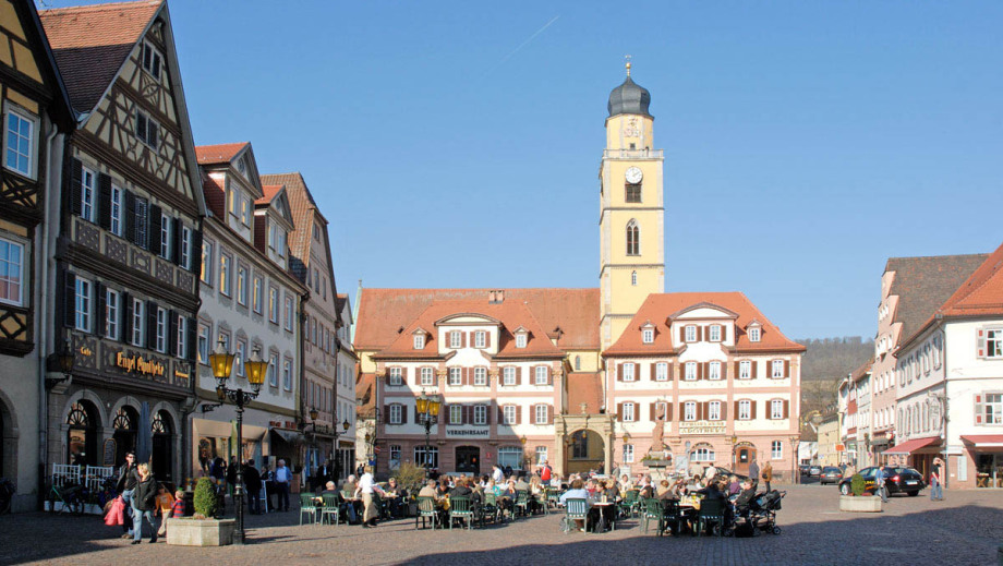 Es ist die Fußgängerzone und die bekannteste Ansicht von Bad Mergentheim. Man blickt auf zwei historische Markthäuser mit einem hohen Turm. Im Vordergrund sitzen viele Menschen an Tischen eines benachbarten Cafés. Alles ist von Sonne beschienen.