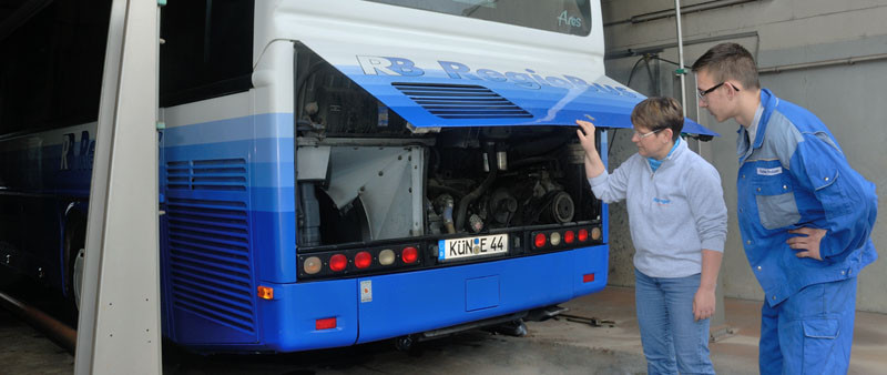 Eine Ausbildungssituation an einem blauen Regiobus der Spedition. Die Heckklappe des Busses ist geöffnet und zwei Mitarbeiter schauen sich den Motor an. Der junge Mitarbeiter hat einen blauen Dress an, die Mitarbeiterin hält die Klappe des Busses.