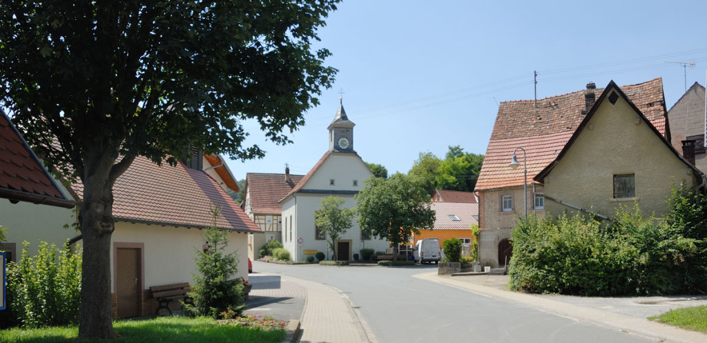 In Epplingen führt eine Straße zur Bildmitte, wo sie sich gabelt. Dort steht ein Haus mit einem kleinen Turm mit Uhr. Rechts und links sind ebenfalls Wohnhäuser. Der Himmel ist blau, die Sonne bescheint die Szene.