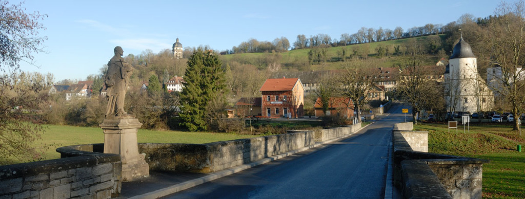 Man fährt auf Schöntal zu und vom Kloster sieht man nur einen kleinen Turm im Bild rechts. Die Straße führt über eine kaum erkennbare Brücke mit einer Heiligenfigur am linken Bildrand. Hinter dem Ortseingangsschild sind weit entfernt Häuser erkennbar.