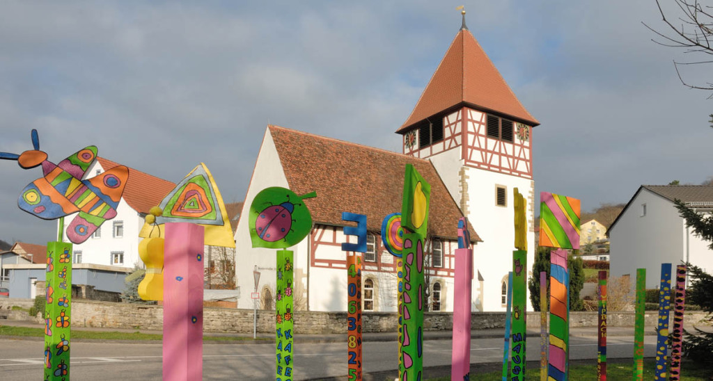 Eine kleine Kirche in Weißbach steht hinter einer Straße. Fachwerkanteile zieren Turm und Schiff des Kirchleins. Davor sind runde zehn Stelen zu sehen: Kunterbunt, von Kindern gemalt. Zwischen Stelen und Kirchlein führt eine Straße waagrecht.