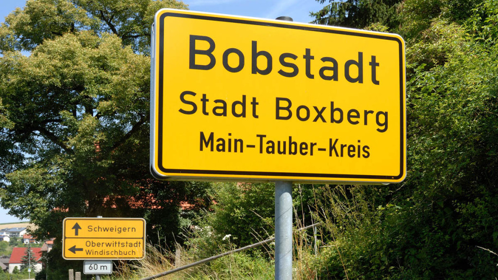 Das Bild zeigt ausschließlich zwei Verkehrsschilder. Das vordere ist das Ortseingangsschild von Bobstadt mit der Addition Stadt Boxberg und dem Hinweis Main-Tauber-Kreis. das hintere Schild weist den Weg. Hinter beiden Schildern ist grünes Baumlaub.