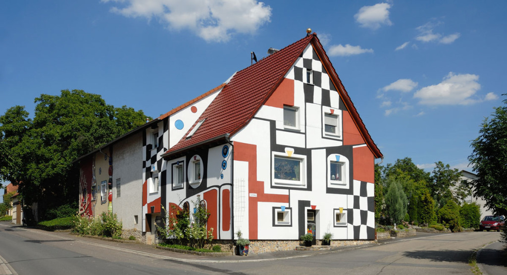 Das wohl bekannteste Haus in Schwabhausen. Es ist ein zweistöckiges Wohnhaus, im Hundertwasserstil bemalt: Es gibt rote Flächen und schwarzweiße im Schachbrettmusterstil. Davor gabelt sich die Straße. Auf beiden Seiten folgt grüne Bepflanzung.