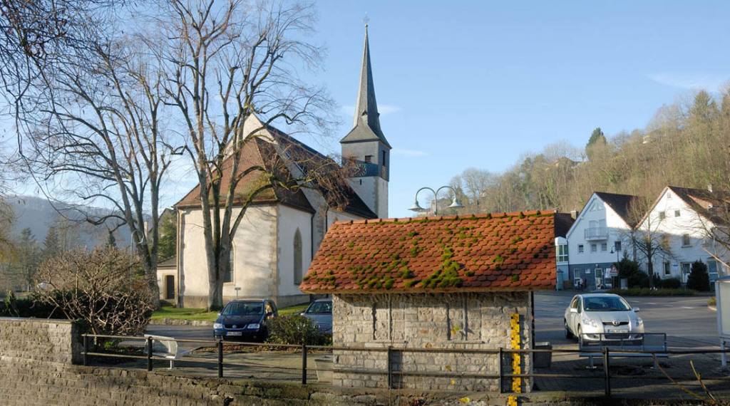 In Widdern sieht man eine Kirche, die hinter einer kleinen mit Ziegeln bedeckten Bushaltestelle steht. rechts sieht man zwei zweistöckige Wohnhäuser. Zwischen Haltestelle und Kirche parken drei Pkw. Zwei große Bäume, links, sind unbelaubt.