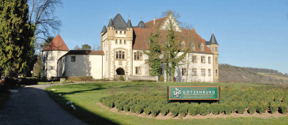 Die Götzenburg, wie man auf einem Schild lesen kann in Jagsthausen. Ein geteerter Weg führt links am Gebäude vorbei. Vorne ist ein Garten angelegt. Man sieht mehrere verschiedene Türme des Schlosses. Der Himmel ist wolkenlos blau.
