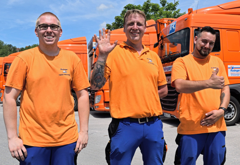 Drei Mitarbeiter haben Spaß beim Fotografieren des Gruppenbildes. Alle drei tragen orangene T-Shirts und blaue Hosen. der mittlere Mitarbeiter winkt dem Fotografen, der recht zeigt „Daumen hoch".